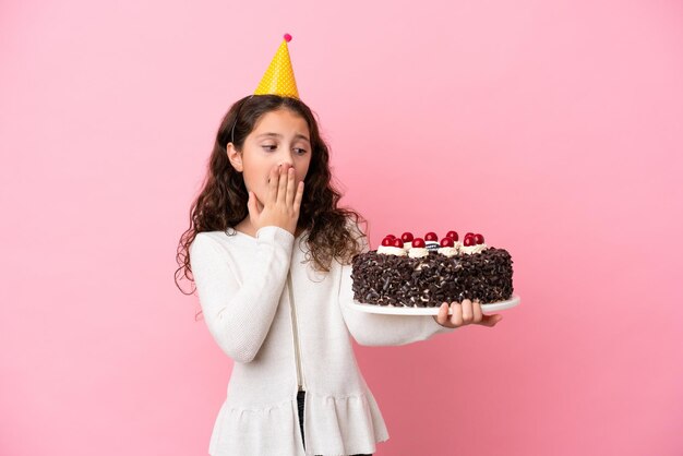 Petite fille caucasienne tenant un gâteau d'anniversaire isolé sur fond rose avec une expression faciale surprise et choquée