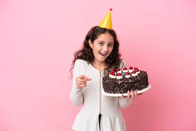 Petite fille caucasienne tenant un gâteau d'anniversaire isolé sur fond rose surpris et pointant vers l'avant