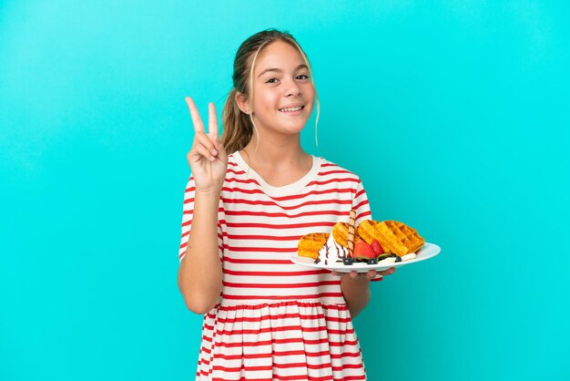 Petite fille caucasienne tenant des gaufres isolées sur fond bleu souriant et montrant le signe de la victoire