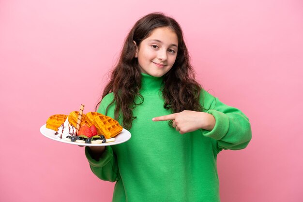 Petite fille caucasienne tenant des gaufres isolées sur fond rose et le pointant