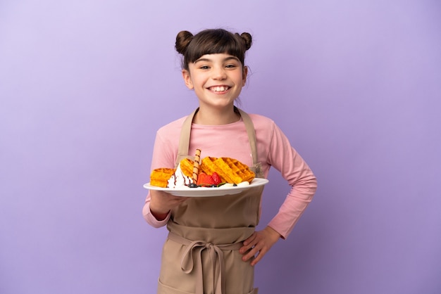 Petite fille caucasienne tenant des gaufres isolées sur un mur violet posant avec les bras à la hanche et souriant