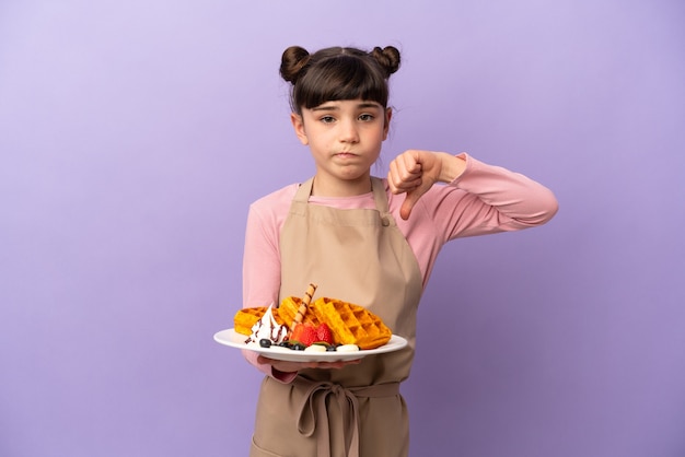 Petite fille caucasienne tenant des gaufres isolées sur violet montrant le pouce vers le bas avec une expression négative