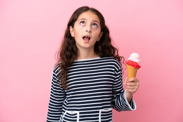 Petite fille caucasienne tenant une glace isolée sur fond rose en levant et avec une expression surprise