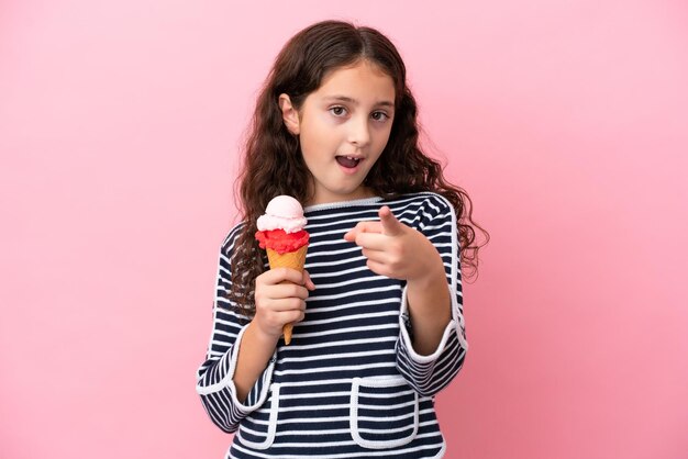 Petite fille caucasienne tenant une glace isolée sur fond rose surprise et pointant vers l'avant