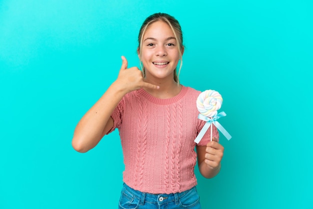 Petite fille caucasienne tenant une sucette isolée sur fond bleu faisant un geste de téléphone. Rappelez-moi signe