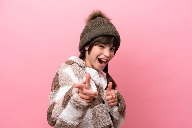 Petite fille caucasienne avec veste d'hiver isolée sur fond rose pointant vers l'avant et souriant