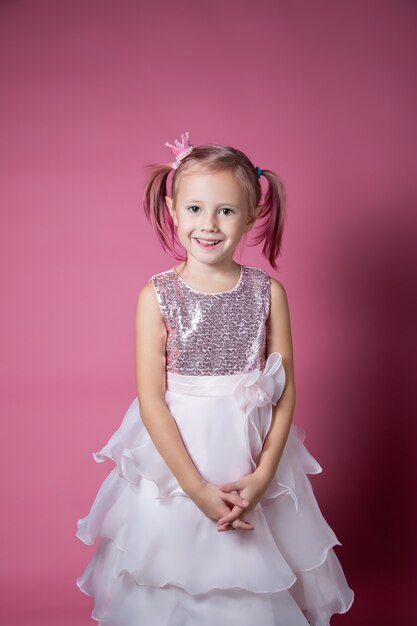 Petite fille caucasienne vêtue d'une robe de fête avec des paillettes posant comme une princesse sur fond rose en regardant la caméra