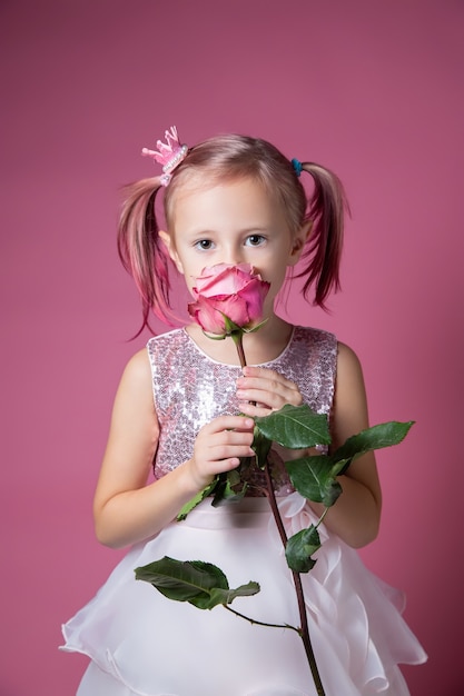 Petite fille caucasienne vêtue d'une robe de fête avec des paillettes posant avec une fleur rose sur fond rose en regardant la caméra