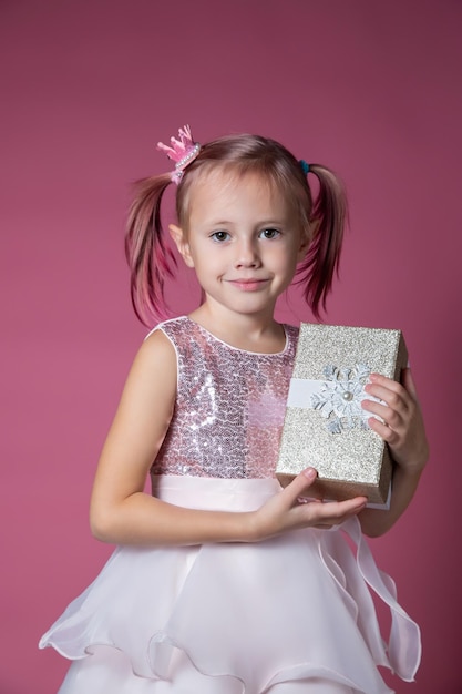 Petite fille caucasienne vêtue d'une robe de fête avec des paillettes posant tenant une boîte-cadeau sur fond rose en regardant la caméra