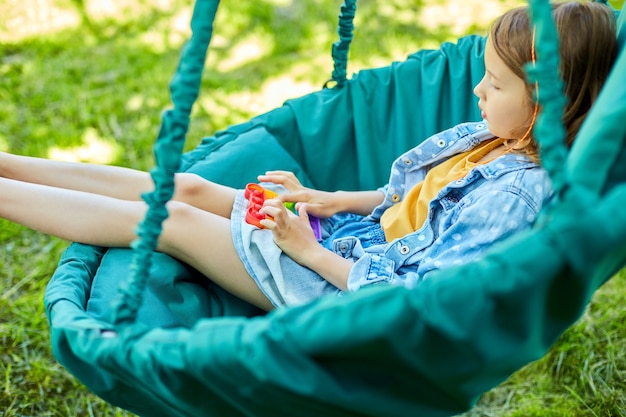 Une petite fille sur une chaise suspendue joue à l'extérieur, des mains d'enfant jouant aux bulles d'un jouet déstressant arc-en-ciel, un jouet agité dans l'arrière-cour de la maison par une journée d'été ensoleillée, vacances d'été