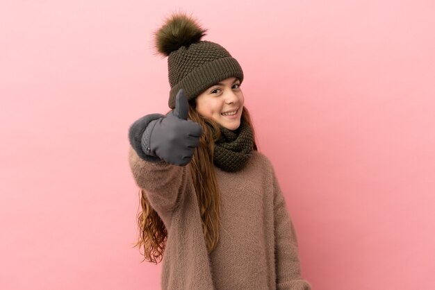 Petite fille avec chapeau d'hiver isolée sur fond rose avec le pouce levé parce que quelque chose de bien s'est produit