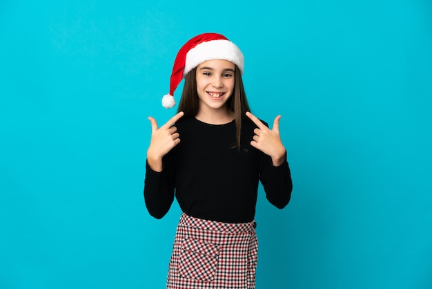 Petite fille avec un chapeau de Noël isolé sur fond bleu donnant un geste de pouce en l'air