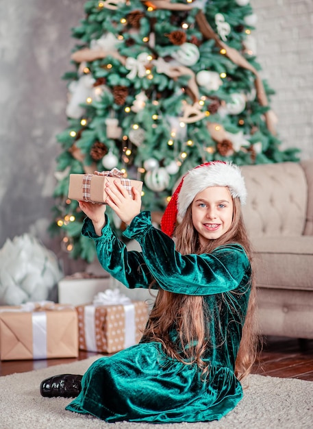 Petite fille chapeau de père Noël avec des cadeaux sous l'arbre de Noël assis près de la cheminée déballe les cadeaux