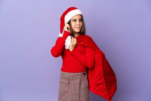 Petite fille avec chapeau et sac de Noël isolé sur mur violet frustré et couvrant les oreilles