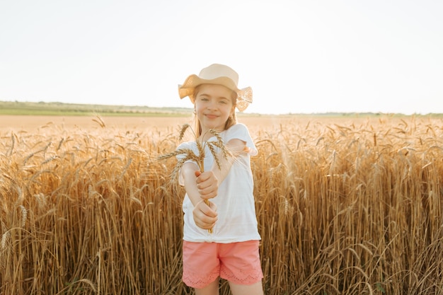 La petite fille avec le chapeau sur la tête sourit et tient des épis de blé doré des champs de blé...