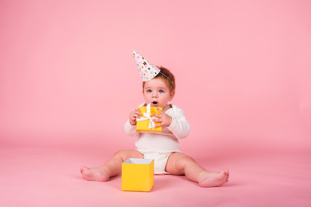 Une petite fille avec un chapeau de vacances est assise avec une boîte jaune sur un mur rose