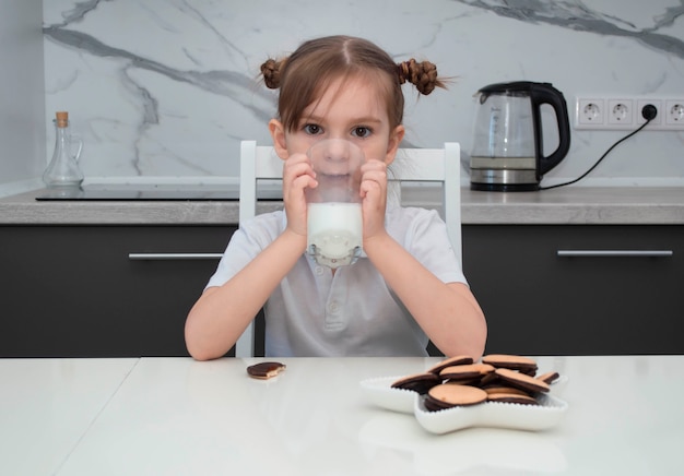 Petite fille charmante en chemise blanche mange des biscuits et boit du lait dans la cuisine de la maison.