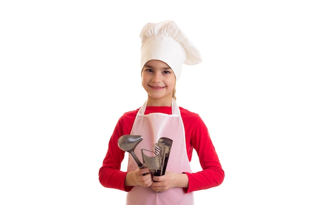 Petite fille charmante en chemise rouge avec tablier blanc et chapeau tenant des ustensiles de cuisine en studio