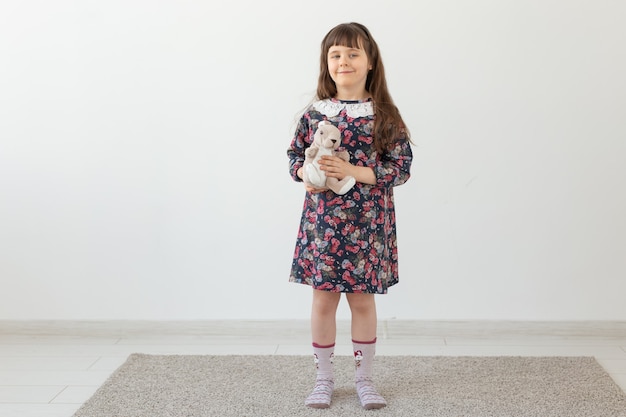 Petite fille charmante dans une robe à fleurs embrasse doucement son lapin jouet préféré debout sur un mur blanc