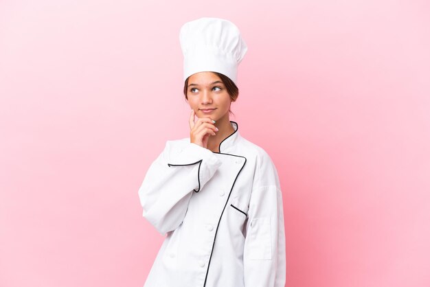 Petite fille de chef caucasien isolée sur fond rose pensant à une idée tout en levant les yeux