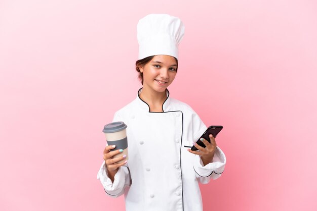 Petite fille de chef caucasien isolée sur fond rose tenant du café à emporter et un mobile