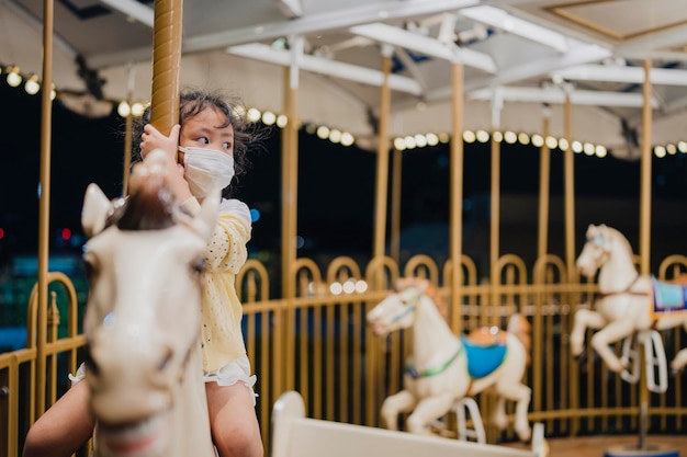 Petite fille sur un cheval de carrousel avec un masque facial sain pour protéger le virus et la pollution.