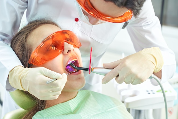 Petite fille chez le médecin. Le dentiste place un remplissage sur une dent avec une lampe de polymérisation dentaire dans la cavité buccale. sur fond de clinique