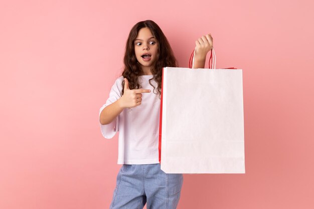 Petite fille choquée pointant sur des sacs à provisions regardant la caméra avec une expression faciale étonnée