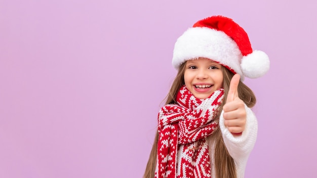 Une petite fille coiffée d'un chapeau du Nouvel An et d'une écharpe chaude est très joyeuse et montre son pouce levé. Un enfant en costume de Noël sur un fond isolé.