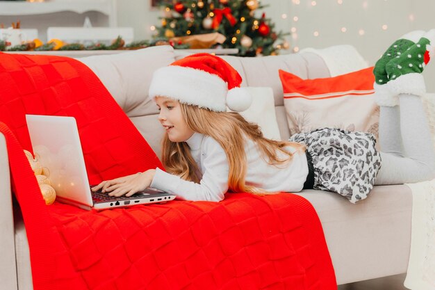 Une petite fille coiffée d'un chapeau de Père Noël communique par appel vidéo via un ordinateur portable, allongée sur le canapé du salon.