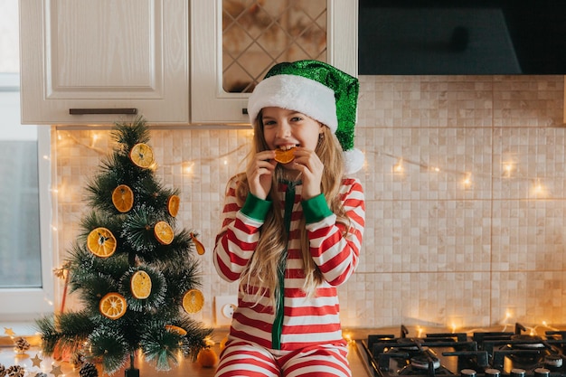 Une petite fille coiffée d'un chapeau de père Noël décore l'arbre de moitiés d'orange séchées. Un bébé mignon se prépare à fêter Noël.
