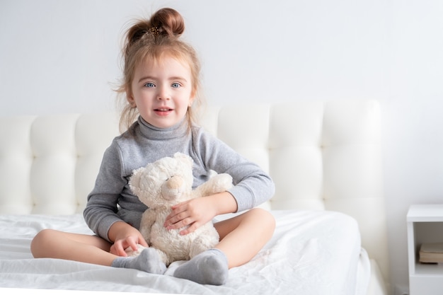 Petite fille en col roulé gris avec ours en peluche assis sur le lit en souriant.