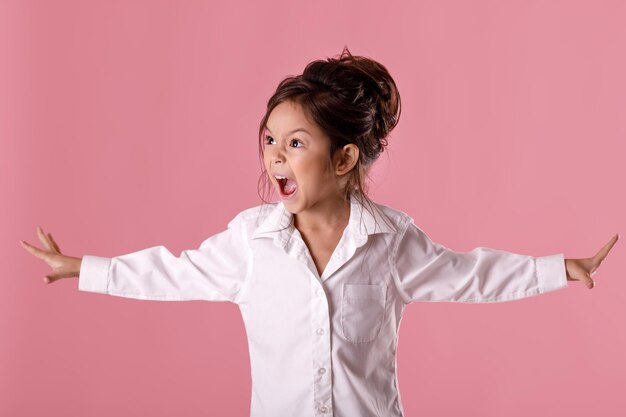 Petite fille en colère qui crie en chemise blanche avec une coiffure en détournant les yeux sur fond rose. Émotions humaines et expression faciale