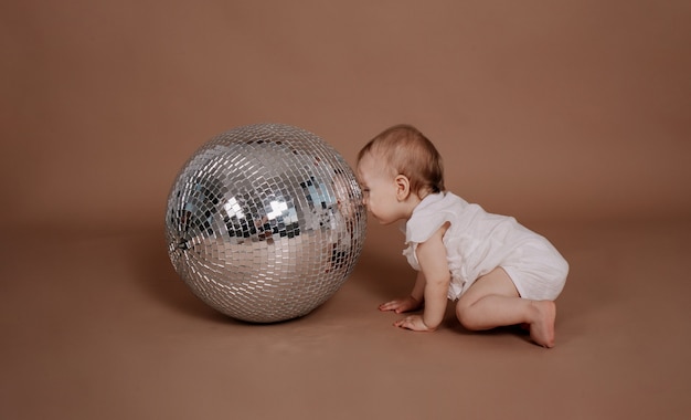 Une petite fille en combinaison blanche regarde une boule disco argentée sur fond beige avec une place pour le texte