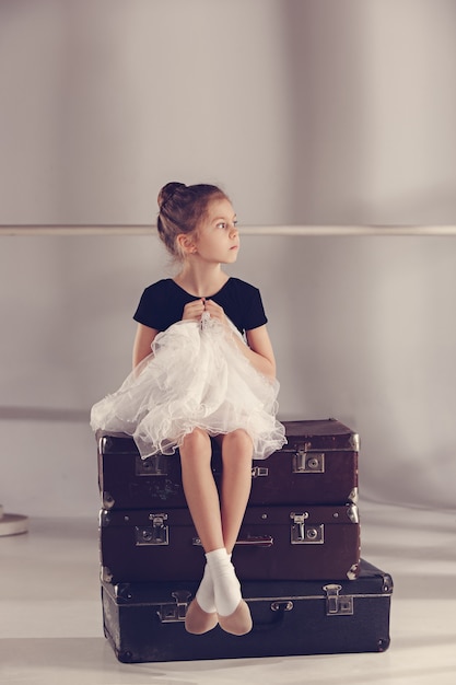 La petite fille comme danseuse balerine assise au studio sur des valises sur fond gris. Les rêves sur les concepts de ballet