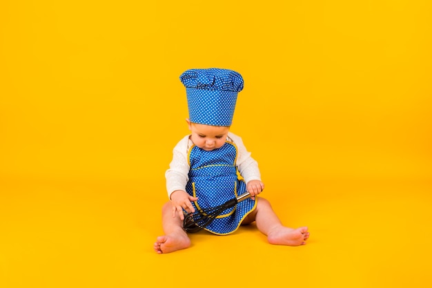 Une petite fille concentrée est assise dans un costume de chef avec un fouet sur un mur jaune avec un espace pour le texte