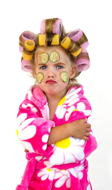 Photo petite fille avec des concombres sur son visage. fille en bigoudis, robe petite coquette.