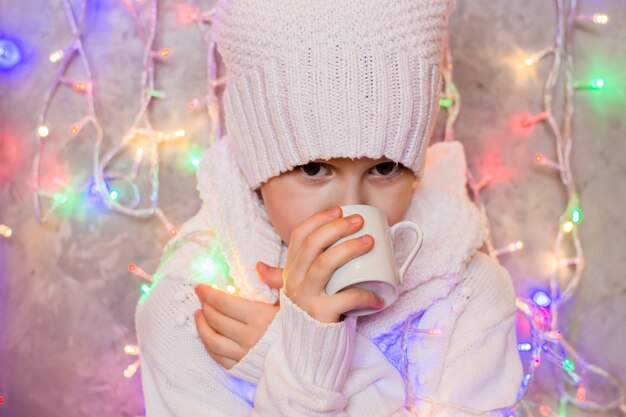 Une petite fille congelée dans un pull et un bonnet blancs boit dans une tasse une boisson chaude sur fond de guirlande colorée. Le concept de froid et de réchauffement