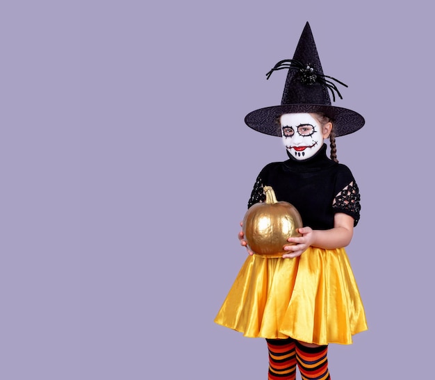 Une petite fille en costume et chapeau d'Halloween tient une citrouille dans ses mains.