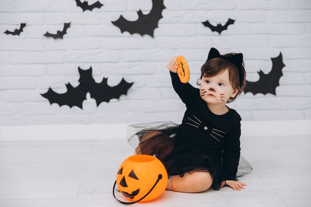 Une petite fille en costume de chat noir avec un panier de citrouille tenant un pain d'épice