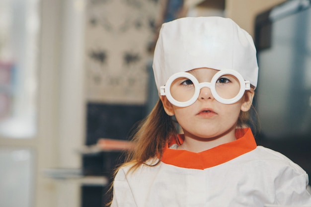 Une petite fille en costume de médecin. L'enfant joue au docteur. Blouse blanche, lunettes, casquette avec une croix rouge, équipement médical. Concept d'un mode de vie sain, soins de santé, médecine