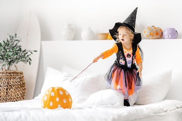 petite fille en costume de sorcière avec baguette magique et citrouille d'Halloween sur le lit à l'intérieur blanc.