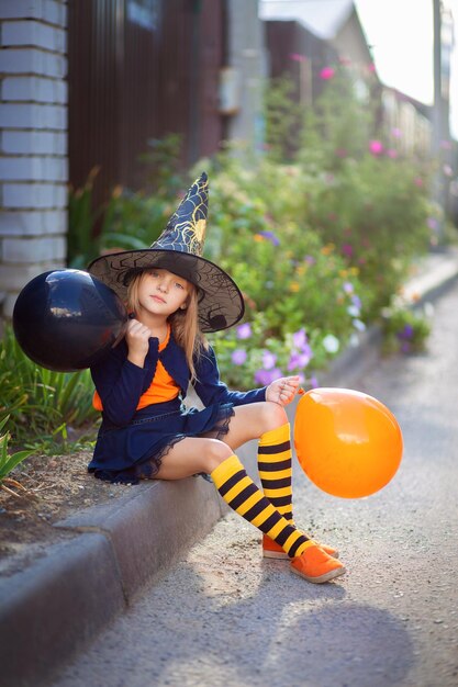 Une petite fille en costume de sorcière et avec des ballons pour Halloween se promène dans la rue et se réjouit des vacances