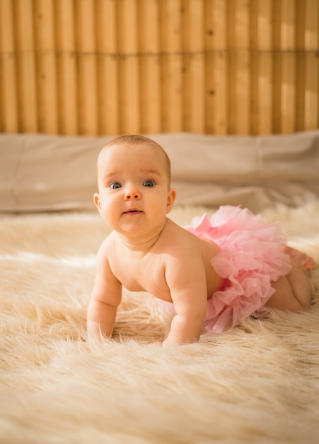 Une petite fille en culotte à volants rose rampe sur une couverture de fourrure dans la chambre