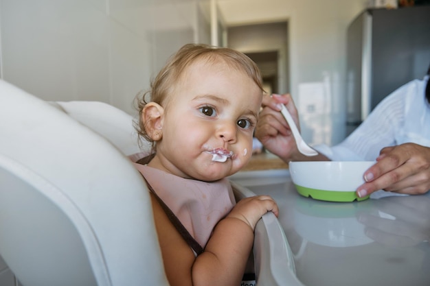 Une petite fille curieuse assise sur une chaise haute et regardant la caméra avec intérêt tout en mangeant avec l'aide d'une femme de récolte dans la cuisine à la maison