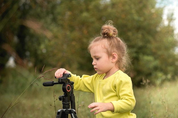 Petite fille curieuse aux cheveux blonds ondulés joue avec un trépied pour des photos dans le parc au chaud