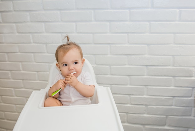 petite fille dans un body blanc est assise sur une chaise d'alimentation avec une cuillère