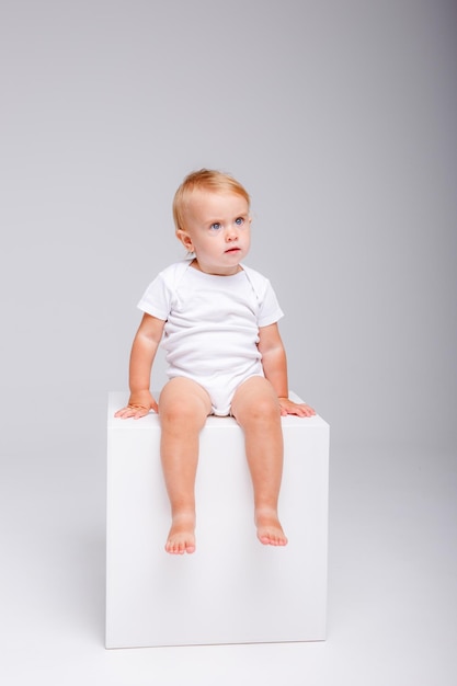 une petite fille dans un body blanc est assise sur un cube blanc sur fond blanc