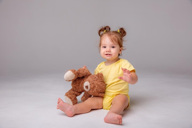 Une petite fille dans un body jaune est assise et joue avec un ours en peluche sur un fond blanc