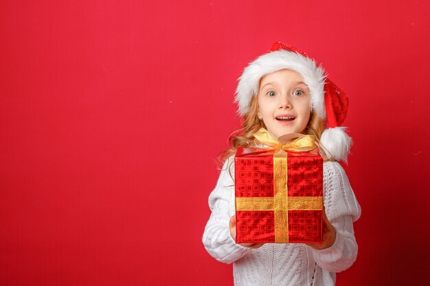 Petite fille dans un bonnet de Noel sur fond rouge avec un gâteau et présent